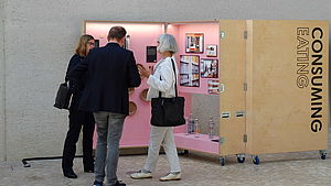Three people standing in front of an exhibition box named Consuming Eating | EcoDesign Circle | Interreg Baltic Sea Region | “Consuming-eating” displays bowls, cups and plates made of bark, wheat bran or even coffee grounds. They showcase sustainable tableware from Lithuania, Poland and Germany – be it for an exceptional disposable single use or a durable every day ware. (© IDZ | International Design Centre Berlin)