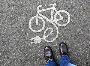 View from above on feet, which stand on the road directly in front of a painted sign: a bicycle with attached power cable | BSR electric - a project of Interreg Baltic Sea Region (photo: Panthermedia/Markus Mainka)