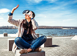 Woman sitting on one of the cuboids on a quay watching her hand through virtual reality glasses | Baltic Game Industry - a project of Interreg Baltic Sea Region (photo: iStock/bedya)