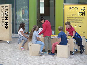 Four children and a grown up sitting on wooden boxes with exhibition boxes in the background | EcoDesign Circle | Interreg Baltic Sea Region | Bringing up the rear, the last box transforms into a table surrounded by wooden seating cubes. At the table, which shows the Baltic Sea region in yellow and black, visitors can pick up an informative exhibition catalogue and sit down on the cubes for a rest, to take it all in, discuss and exchange … (©German Environment Agency | Conrad Dorer)