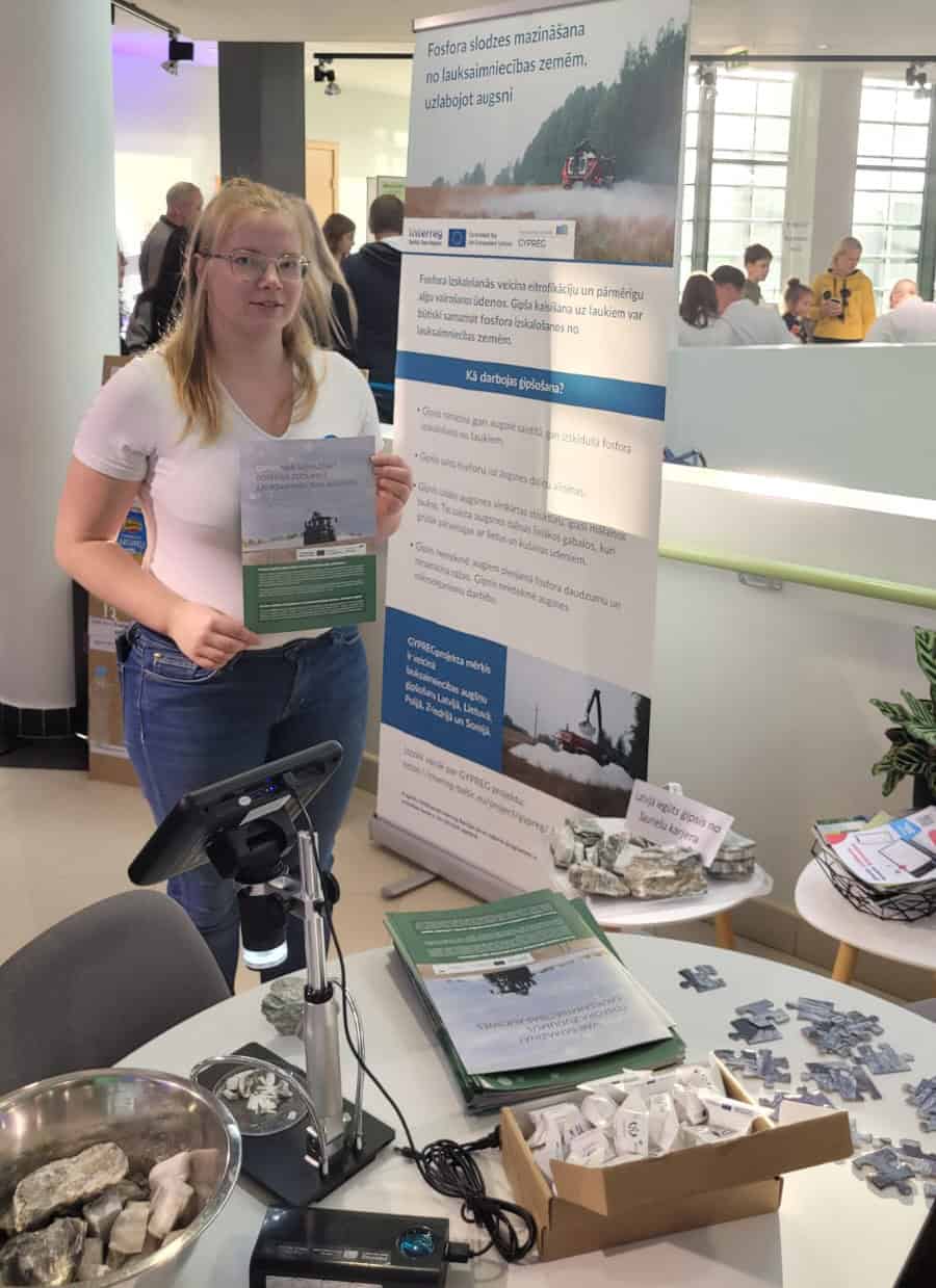 A person presenting the GYPREG brochure at the event. Visitors in the background.