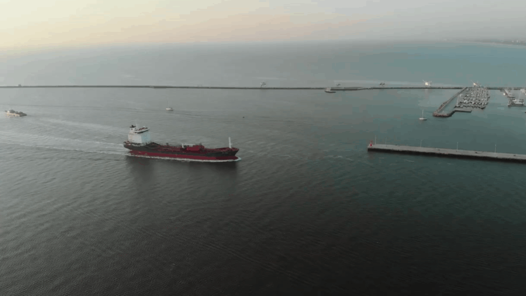 A cargo ship sailing through a harbor channel with breakwaters and a marina visible in the background.