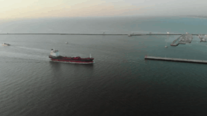 A cargo ship sailing through a harbor channel with breakwaters and a marina visible in the background.